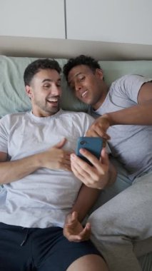 Mutlu çoklu-etnik eşcinsel çift yatakta uzanıyor, sosyal medyada geziniyor, gülüyor ve akıllı telefonu yavaş çekimde kullanıyor.