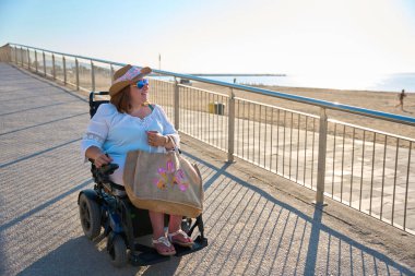 Hasır şapkalı ve güneş gözlüklü kaygısız kadın yaz tatilinin tadını tekerlekli sandalyesiyle çıkarıyor.