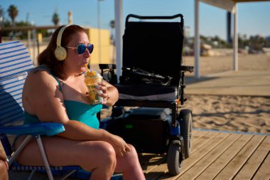 Güneş gözlüklü, kulaklıklı, güneşli bir sahilde meyveli kokteyl içen, tekerlekli sandalyesini yakınlarda park etmiş iri yarı bir kadın.