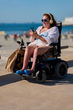Mutlu bir kadın tekerlekli sandalyede güneşli bir günün tadını çıkarırken akıllı telefon kullanıyor ve kulaklıkla müzik dinliyor.
