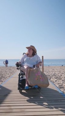 Tekerlekli sandalyedeki kadın tahta kaldırımda geziniyor, güneşli yaz gününde özgürlük ve bağımsızlıkla plaj tatiline çıkıyor.