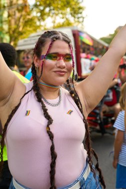 Gurur geçidinde kutlama yapan genç bir kadın, neşeli kollarını kaldırırken canlı gökkuşağı makyajı ve bayrak rozetleriyle süslenmişti.