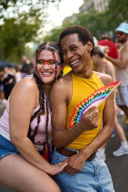 İki insan sarılıp gülüyor, ellerinde gökkuşağı yelpazesi var, bir gurur festivalinin canlı atmosferinin tadını çıkarıyorlar.