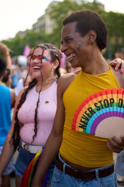 Gurur festivaline katılan iki genç insan birlikte gülüyor. Bir tanesi elinde gökkuşağı hayranı tutuyor.