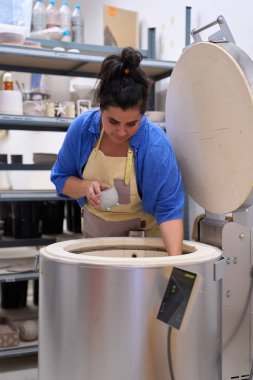 Kadın çömlekçi dikkatlice fırına küçük bir seramik kase yerleştiriyor, onun el yapımı sanatları ile dolu yaratıcı çalışma alanı ile çevrili.