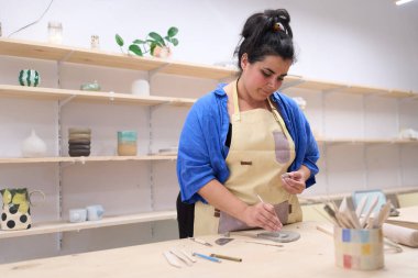 Seramik atölyesinde özenle çalışan kadın zanaatkar, çeşitli aletlerle iyi kullanılmış bir tezgahta killeri şekillendiriyor ve şekillendiriyor.