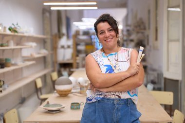 Çanak çömlek atölyesinde gülümseyen, elinde aletlerle hazır bir kadın seramiğin portresi.