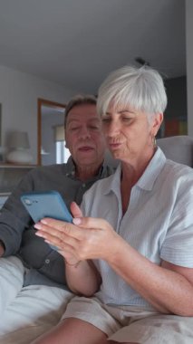 Yaşlı çift, yavaş çekim sekansıyla çekilen bir kanepede birlikte akıllı telefon kullanırken bir anlık eğlencenin tadını çıkarıyor.