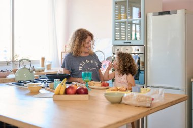 Anne ve kız birlikte kahvaltı yapıyorlar. Parlak, modern mutfaklarında, yemek ve sohbetlerini paylaşıyorlar.