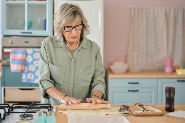 Yaşlı kadın merdane kullanıyor, hamuru mutfak tezgahında düzleştiriyor, kurabiye yapmaya hazırlanıyor.