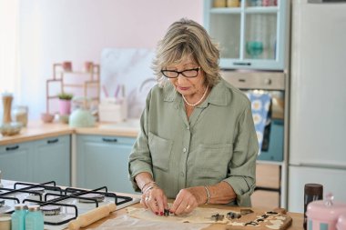 Sıcak ve sevgi dolu, parlak, modern bir mutfakta ev yapımı kurabiyeler hazırlarken yaşlı bir kadın neşe içinde kurabiye keskisi kullanıyor.