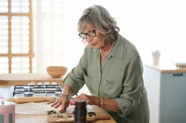 Yaşlı bir kadın, sevgi ve sıcaklıkla dolu, ev yapımı kurabiyeler hazırlarken, neşeli bir şekilde kurabiye keskisi kullanıyor.