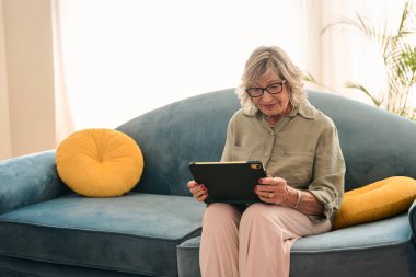 Yaşlı bir kadın mavi bir kanepede rahat bir şekilde oturuyor, dijital tablet kullanıyor, parlak bir oturma odasında modern teknolojinin tadını çıkarıyor.