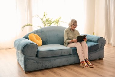 Oturma odasında rahat bir koltukta otururken dijital tablet kullanan yaşlı bir kadın modern teknolojinin tadını çıkarıyor.