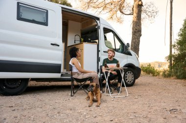 Genç çift karavanın dışında oturmuş, köpekleriyle içki içip sohbet ediyorlar.