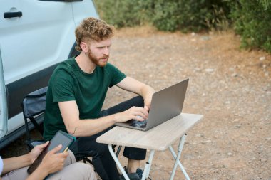 Genç adam, karavanının yanındaki kamp koltuğunda oturup dijital göçebe yaşam tarzının keyfini çıkarırken uzaktan bilgisayarında çalışıyor.