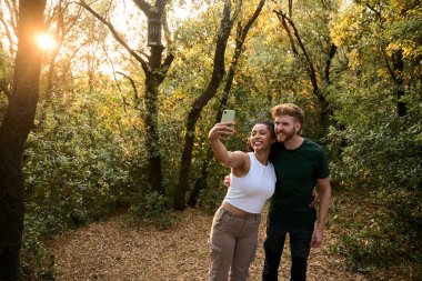 Mutlu çift ormanda günbatımı yürüyüşünün tadını çıkarıyor, güzel doğanın ortasında akıllı telefonlarıyla neşeli bir selfie çekiyorlar.