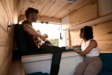 Kız arkadaşı için akustik gitar çalan genç bir adam yolculuk sırasında rahat bir karavanda dinleniyor.