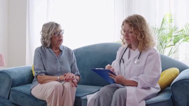 Ev ziyareti sırasında semptomları tarif eden yaşlı hastayı dinlerken not alan kadın doktor