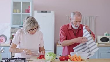 Kıdemli çift birlikte yemek hazırlıyor, sebze doğruyor ve parlak bir mutfakta ellerini siliyor. Yavaş çekim
