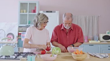 Olgun çift, sağlıklı bir yemek hazırlarken birlikte iş birliği yapıyor, güneş ışığında taze sebzeleri dikkatlice kesiyor, yavaşça ve kasten hareket ediyor.