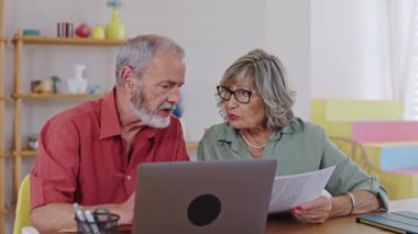 Yaşlı çift belgeleri inceliyor ve ağır çekimde yakalanan dizüstü bilgisayarla tartışıyor.
