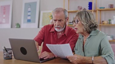 Yaşlı çift evrakları inceliyor ve dizüstü bilgisayar kullanıyor, yavaş çekimde, ifadelerini ve hareketlerini vurguluyor.
