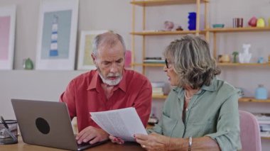 Üst düzey çift finansal belgeleri dikkatlice gözden geçiriyor ve birlikte dizüstü bilgisayarda geziniyor, ev ortamında ağır çekimde çözülen işbirlikçi problem çözme görüntülerini yakalıyorlar.