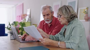 Yaşlı çift evrakları inceliyor ve dizüstü bilgisayar kullanıyor. Endişelerini dile getiriyor ve önemli konuları ağır çekimde tartışıyor.