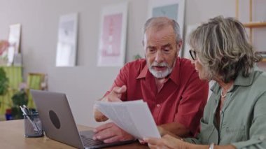 Yaşlı çift evrakları inceliyor ve dizüstü bilgisayar kullanıyor. Yavaş çekimde bilgiyi tartışıyor ve işaret ediyor.