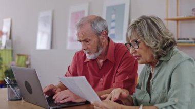 Yaşlı çift evrakları inceliyor ve dizüstü bilgisayar kullanıyor. Yavaş çekimde bilgileri birlikte analiz ediyorlar.