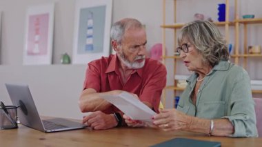 Finansal gerilim yaşayan yaşlı ortaklar, ev giderlerini artan bir hayal kırıklığıyla değerlendirirken masada oturuyor ve yavaş çekimde artan ekonomik baskıları tartışıyorlar.