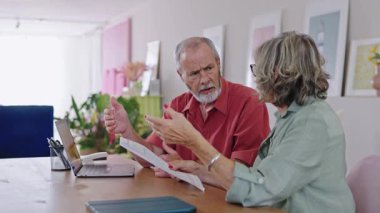 Yaşlı bir çift, elinde dizüstü bilgisayarla masada oturarak evrak işleriyle, muhtemelen faturalarla veya tıbbi sonuçlarla ilgilenerek tepki veriyor.