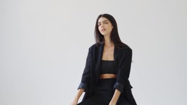 Moda fotoğraflarında çeşitli pozlar veren profesyonel bir model, siyah bir takım elbise giyiyor, beyaz bir arka planda, yavaş çekimde izole edilmiş.