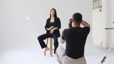 Profesyonel fotoğrafçı, beyaz arka planda, ağır çekimde bir stüdyoda taburede poz veren bir modelin fotoğraflarını çekiyor.