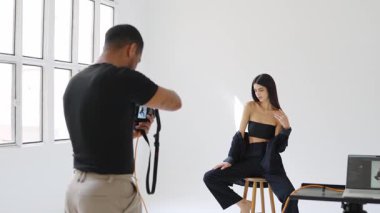 Fotoğrafçı, beyaz arka planda, yavaş çekimde, bir stüdyoda taburede poz veren mankenin fotoğraflarını çekiyor.