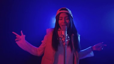 Genç bayan şarkıcı, günlük giysiler ve beysbol şapkası giyerek tutkuyla şarkı söylüyor, canlı, renkli bir ortamda bir mikrofona şarkı söylüyor.