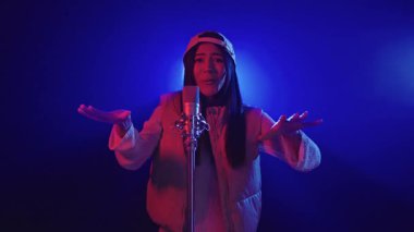 Bej yelek ve beyzbol şapkası giyen enerjik genç bayan şarkıcı mavi ve kırmızı sahne ışıkları altında ağır çekimde tutkuyla performans sergiliyor.