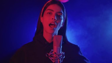 Genç bir kadın bir kayıt stüdyosunda mikrofonla şarkı söylüyor, mavi ve kırmızı ışıklarla aydınlatılıyor, yavaş çekimde.