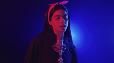 Genç bir kadın bir kayıt stüdyosunda mikrofonla şarkı söylüyor. Renkli ışıklarla aydınlatılıyor. Yavaş çekim.