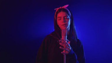 Mavi ve kırmızı ışıklarla aydınlatılmış, mikrofonla şarkı söyleyen genç bir kadının yavaş çekim videosu.