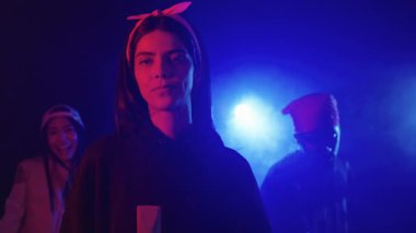 Dumanlı, neon ışıklı bir ortamda arkadaşlarıyla şarkı söyleyen kendine güvenen genç bayan şarkıcı yavaş çekimde yakalanır.