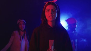 Genç bir kadının mikrofonla şarkı söylemesini, grup arkadaşlarının dumanlı, neon ışıklı bir ortamda dans edip enstrüman çalmasını gösteren yavaş çekim görüntüleri.