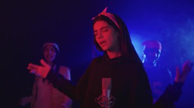 Şarkıcı, renkli spot ışıklarıyla aydınlatılmış bir stüdyoda arka planda dansçılarla şarkı söylüyor.