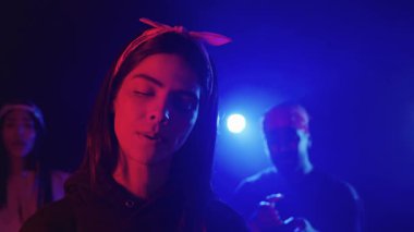 Sahnede tutkulu bir şekilde şarkı söyleyen genç bir kadının yavaş çekim montajı, mavi ve kırmızı ışıklarla aydınlatılmış, arka planda geri vokaller ve müzisyenler var.