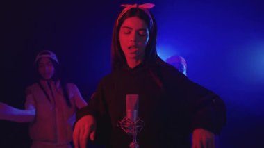 Mavi ve kırmızı ışıklarla aydınlatılmış karanlık bir stüdyoda grubuyla birlikte şarkı söyleyen genç bir kadının yavaş çekim videosu.