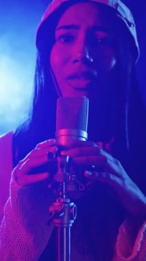 Mavi ve kırmızı ışıklarla aydınlatılmış bir kayıt stüdyosunda mikrofonla şarkı söyleyen genç bir kadın şarkıcının yavaş çekim montajını kapatın.