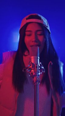 Yetenekli genç bayan şarkıcı, beysbol şapkası takıyor sahnede tutkuyla şarkı söylüyor, parlak mavi ve kırmızı ışıklarla aydınlatılıyor.