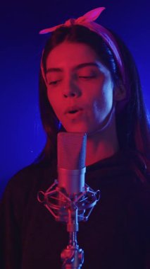 Genç bir kadın bir kayıt stüdyosunda şarkı söylüyor. Mavi ve kırmızı ışıklarla aydınlatılıyor. Ağır çekimde.
