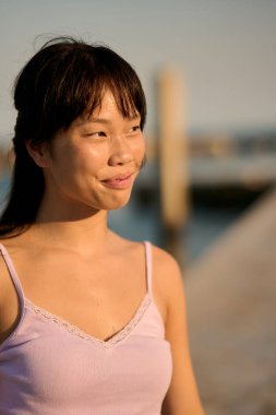 Genç Asyalı bir kadının portresi mutlu bir şekilde gülümseyerek uzaklara bakıyor, sakin bir akşamda altın saatin sıcacık ışığında güneşleniyor.